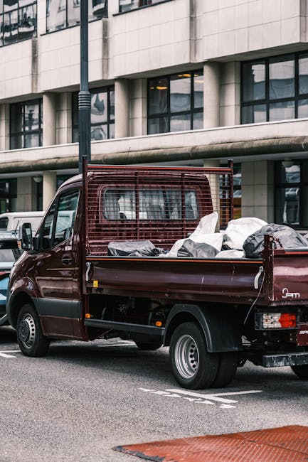 A small, dark red flatbed truck parked on an urban street in front of a modern building with large glass windows and a concrete facade. The truck's cargo area is filled with various types of waste mat