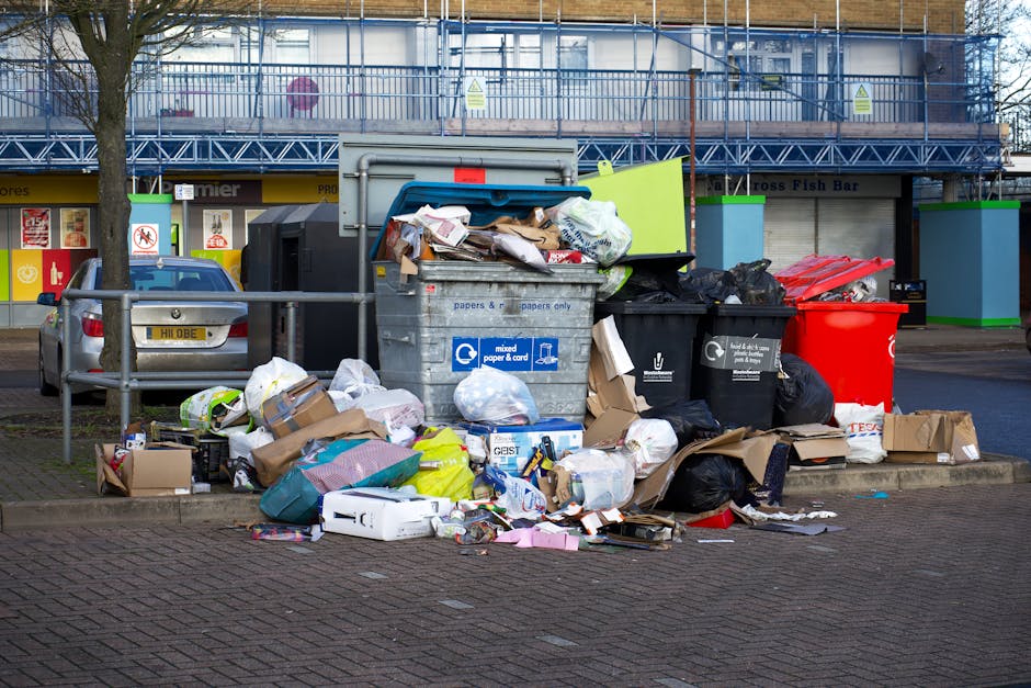 A municipal waste collection worker wearing a red and yellow uniform is standing at the rear of a large, red rubbish collection truck with its rear hatch open, positioned on a street. The worker is lo