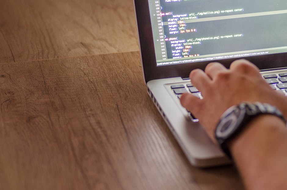 A close-up view of a person's right hand typing on a silver laptop keyboard, which is placed on a wooden desk with a natural grain finish. The laptop screen displays lines of code with a dark backgrou